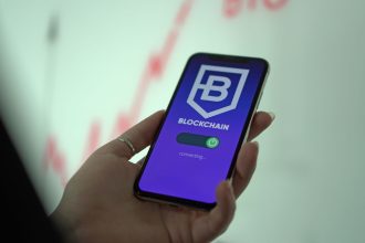 Mão segurando um smartphone que exibe uma carteira digital de criptomoedas e elementos de blockchain